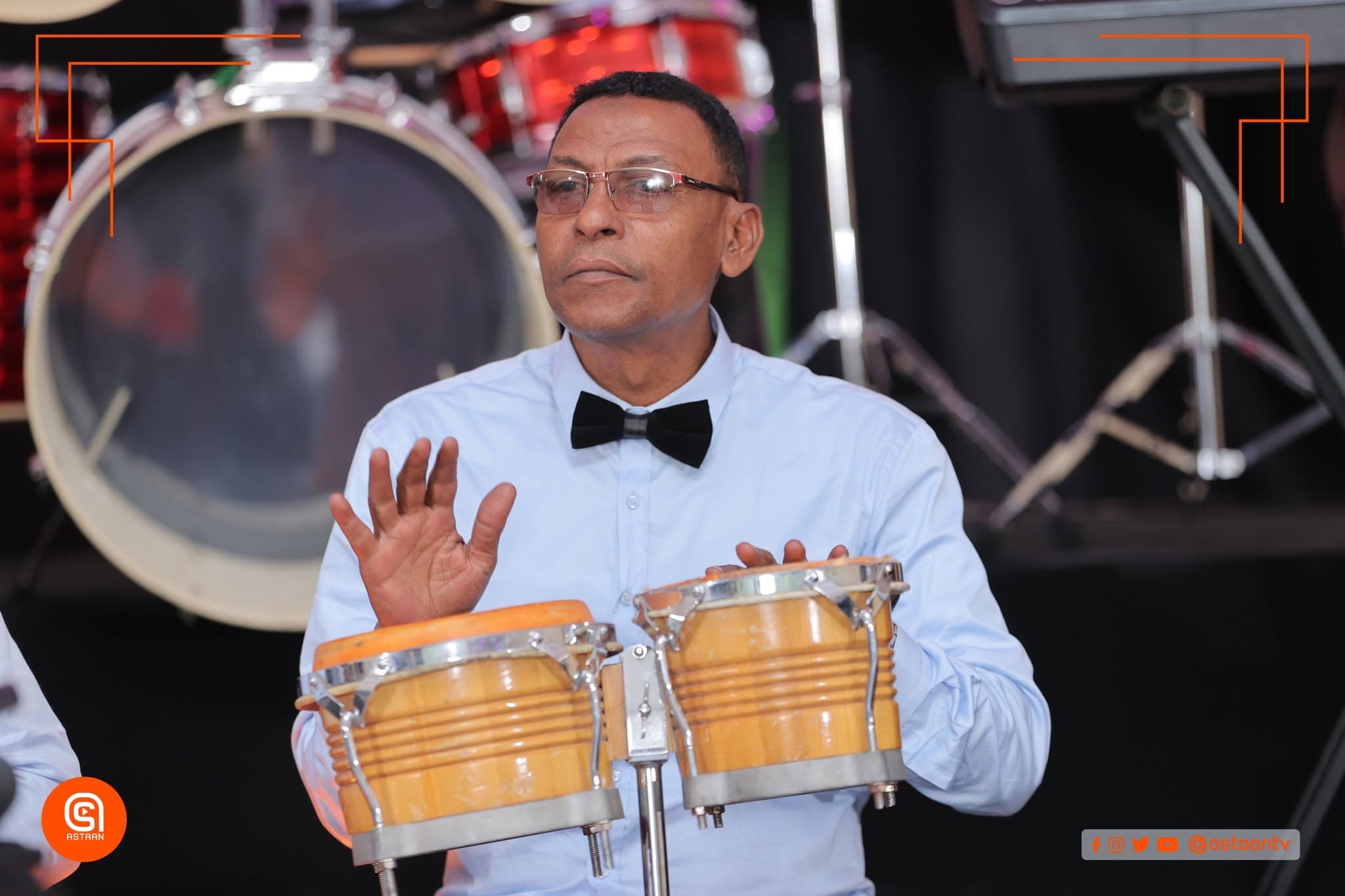 A percussionist with glasses, white shirt and black bow tie playing bongos, a drum kit behind him.