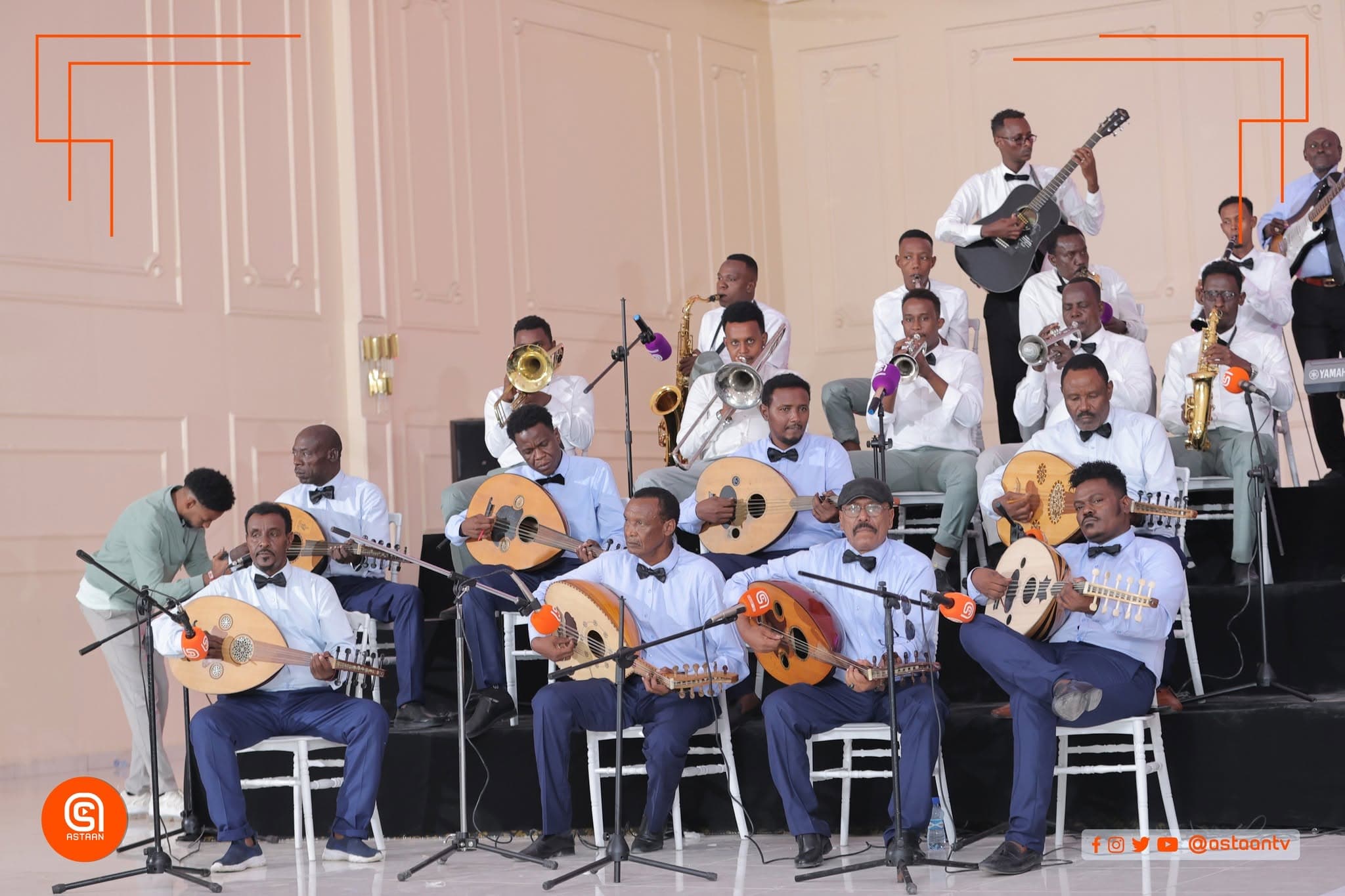 The ensemble seated in rows against a pale panelled wall — a front line of oud players in white shirts and bow ties, with saxophone, trombone and trumpet players and a guitarist behind.