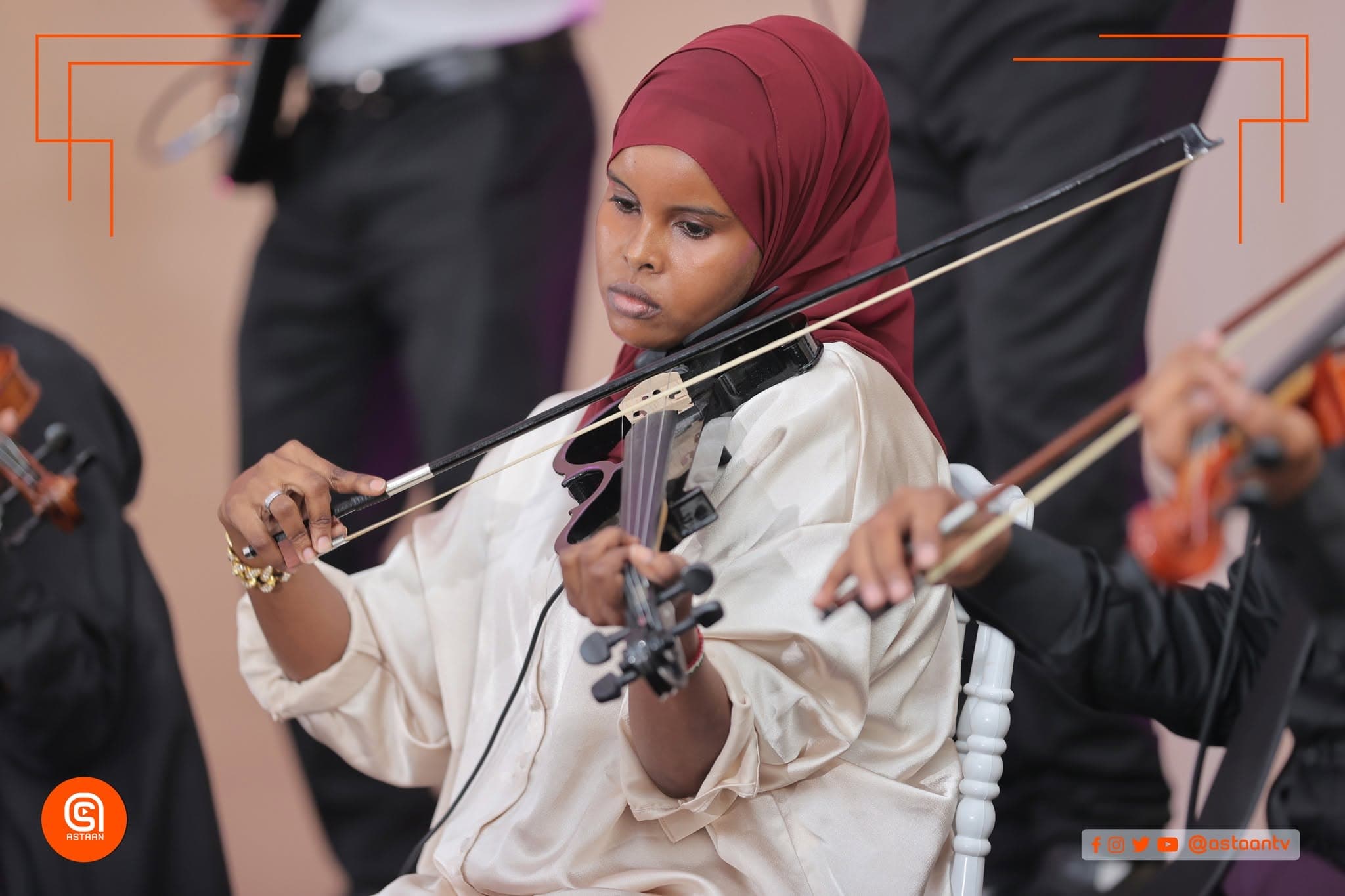 A young violinist in a maroon hijab and cream blouse drawing her bow across the strings, another violinist playing beside her.