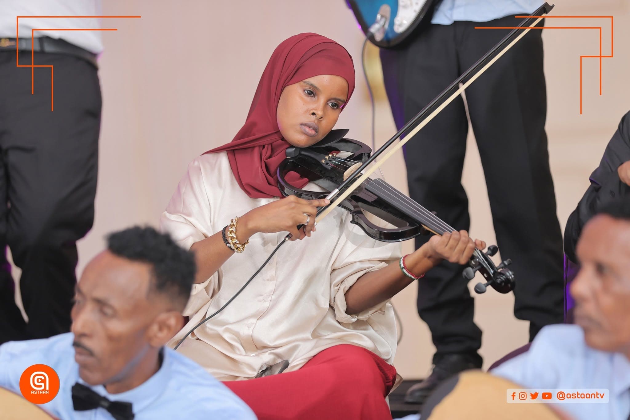 A violinist in a maroon hijab playing an electric violin while seated among fellow musicians in bow ties.