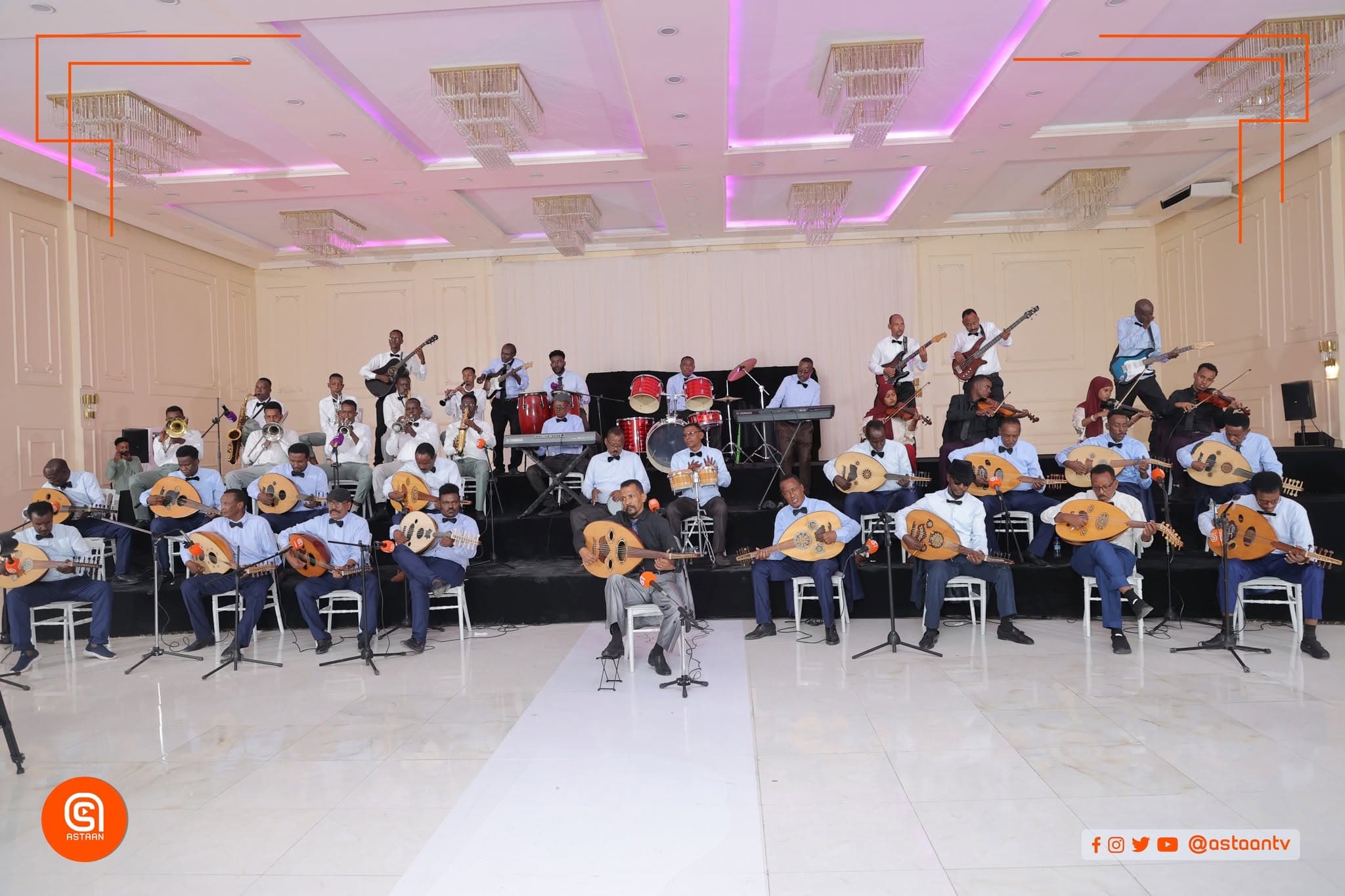 Wide view of the 41-piece orchestra performing beneath chandeliers and magenta ceiling lights — seated oud players in front, drums, keyboards, guitarists and violinists on risers behind.