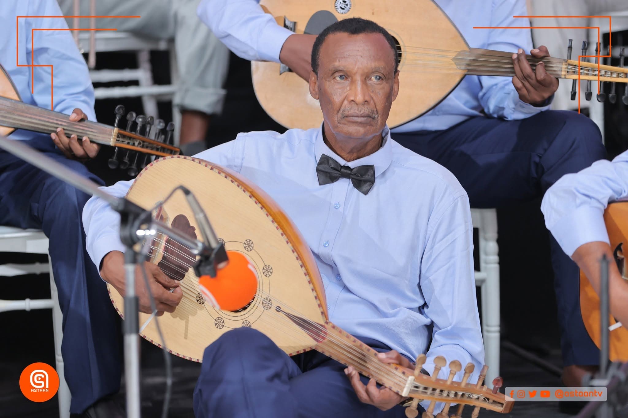 A veteran oud player in a light-blue shirt and black bow tie seated mid-performance, surrounded by fellow oud players.