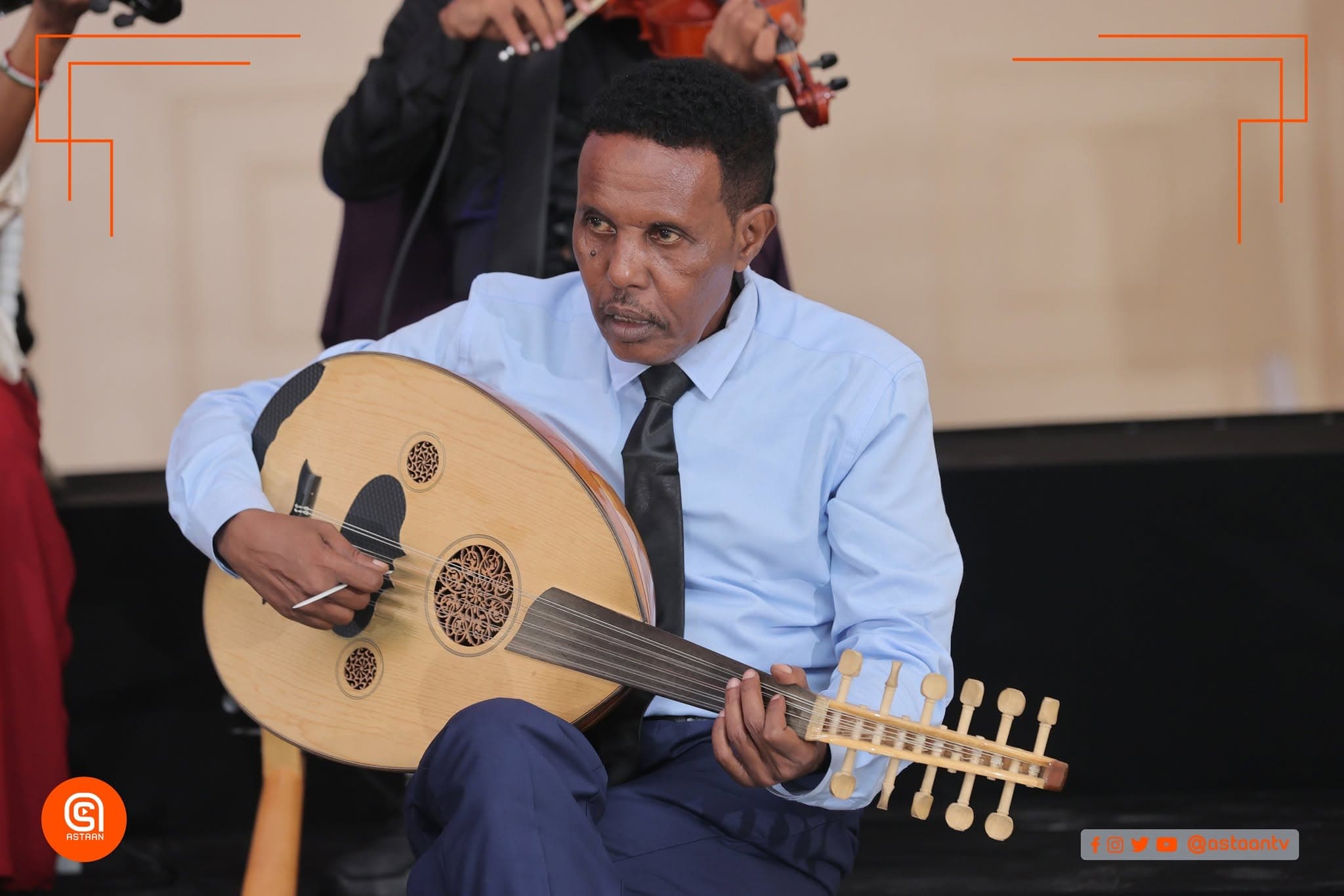 Close view of a musician in a blue shirt and tie playing an intricately decorated oud, with violinists standing behind.