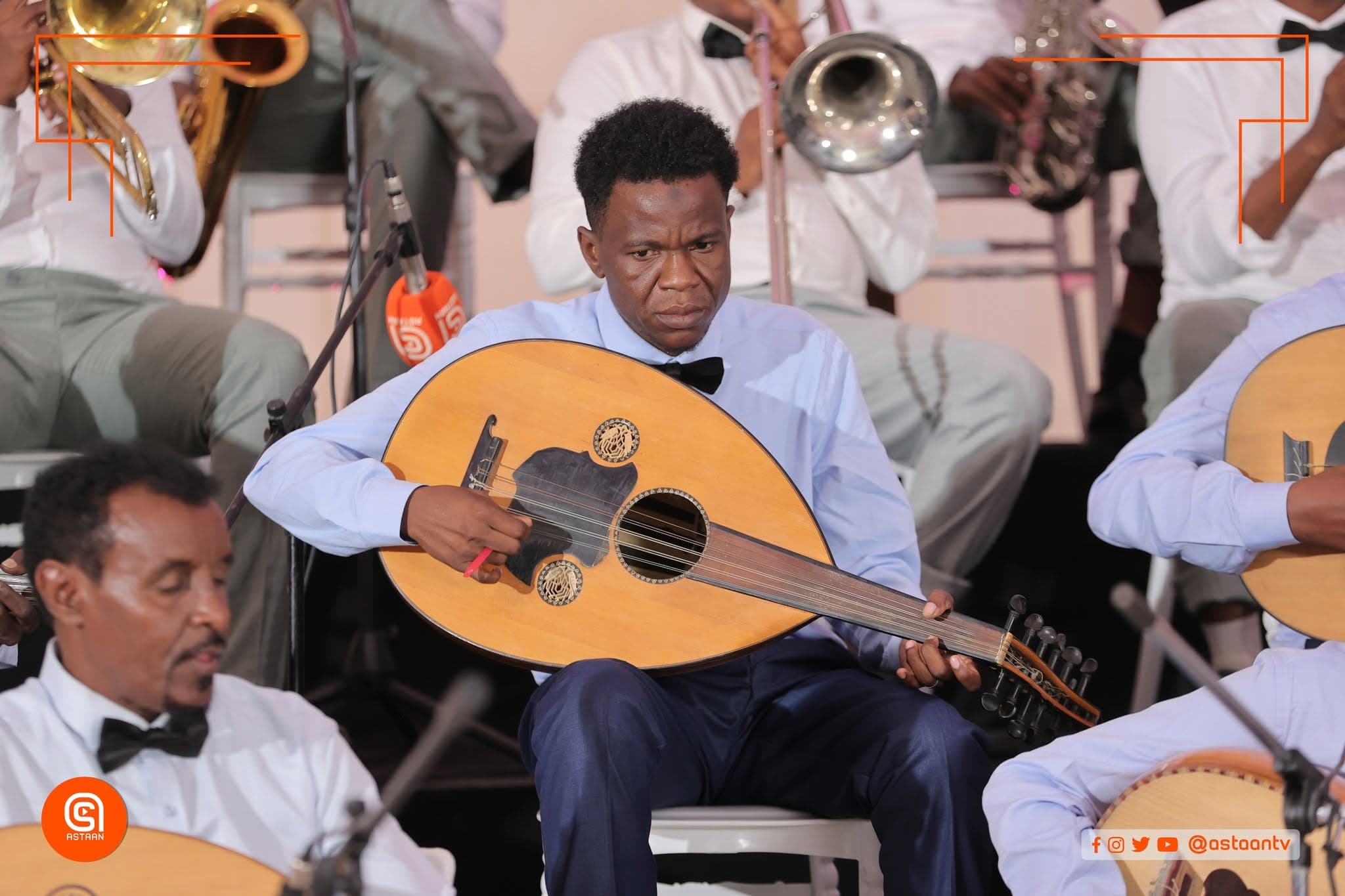 An oud player in a blue shirt and black bow tie mid-song, brass and saxophone players seated behind him.