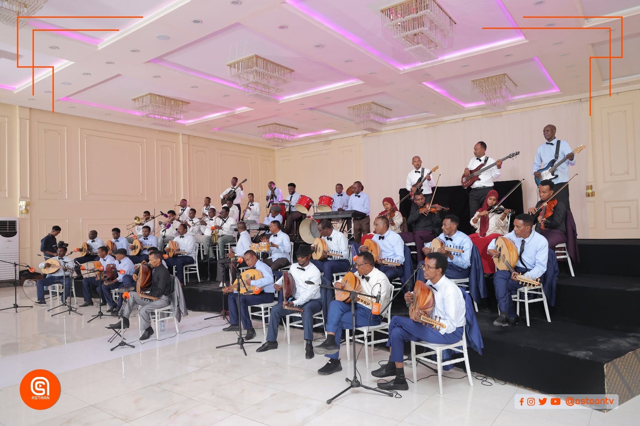 The Hargeisa ensemble in a white hall with purple ceiling lights — seated oud players in light-blue shirts in front, guitarists, drummers and violinists arranged on the platform behind.