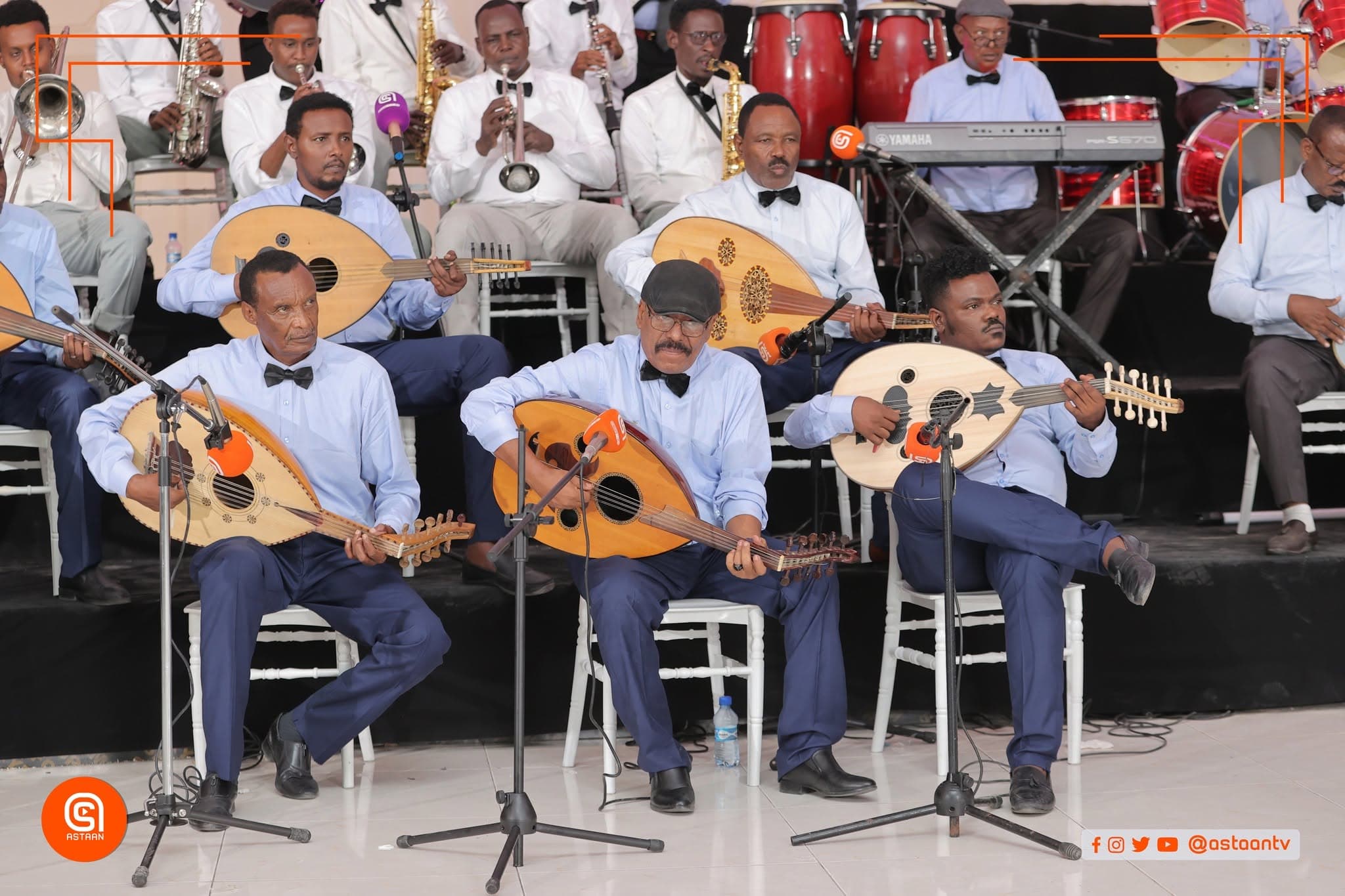 Three seated oud players performing side by side, with red congas and a drum kit on the platform behind them.
