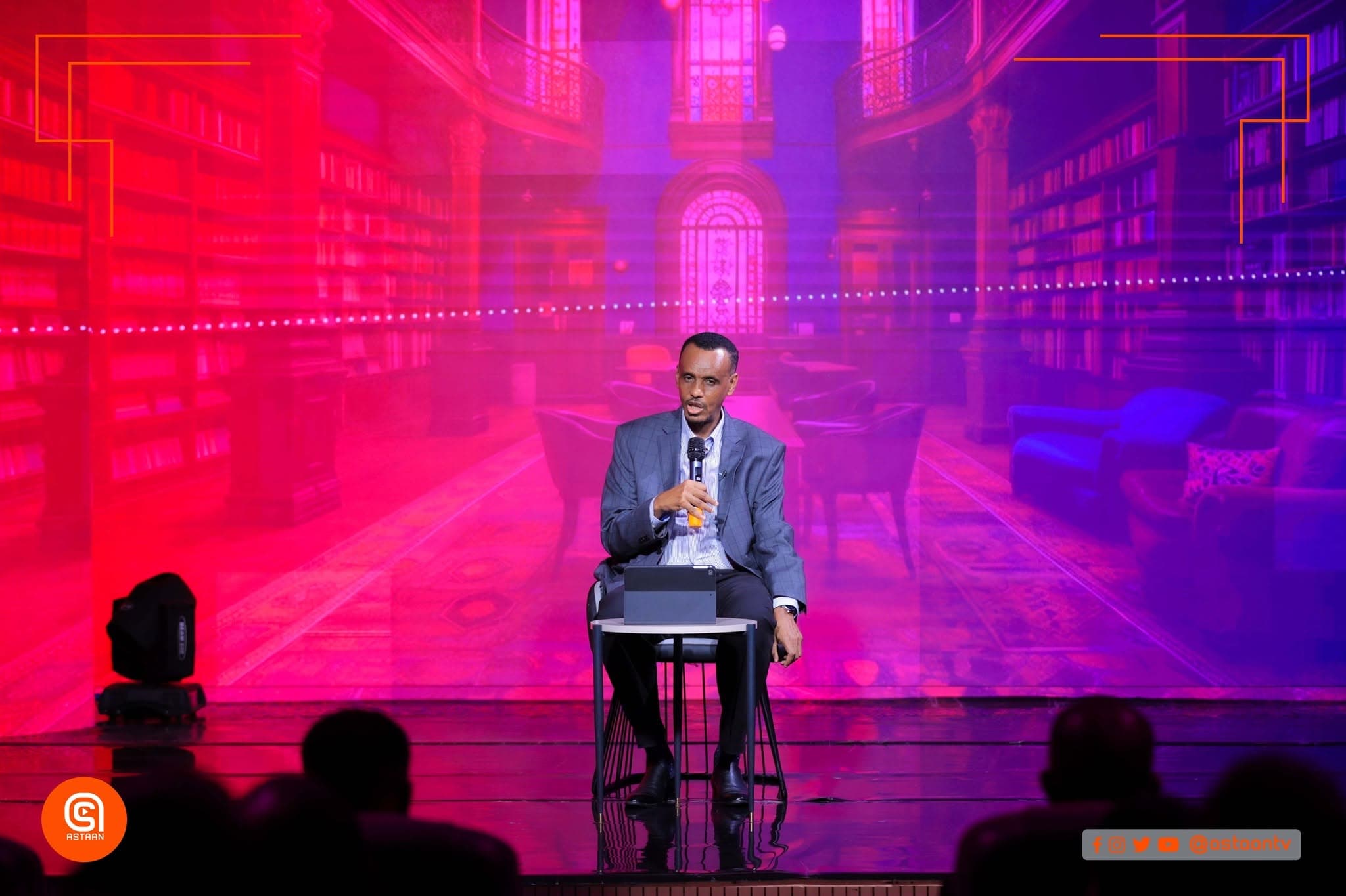 Speaker in a gray suit seated on stage with a microphone under dramatic red and pink lighting, a virtual library glowing on the LED wall