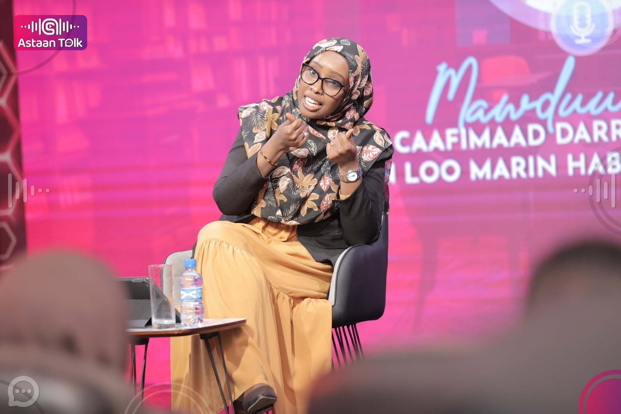 Guest speaker in a floral headscarf gesturing mid-discussion on stage, framed by a magenta LED backdrop with a Somali session title