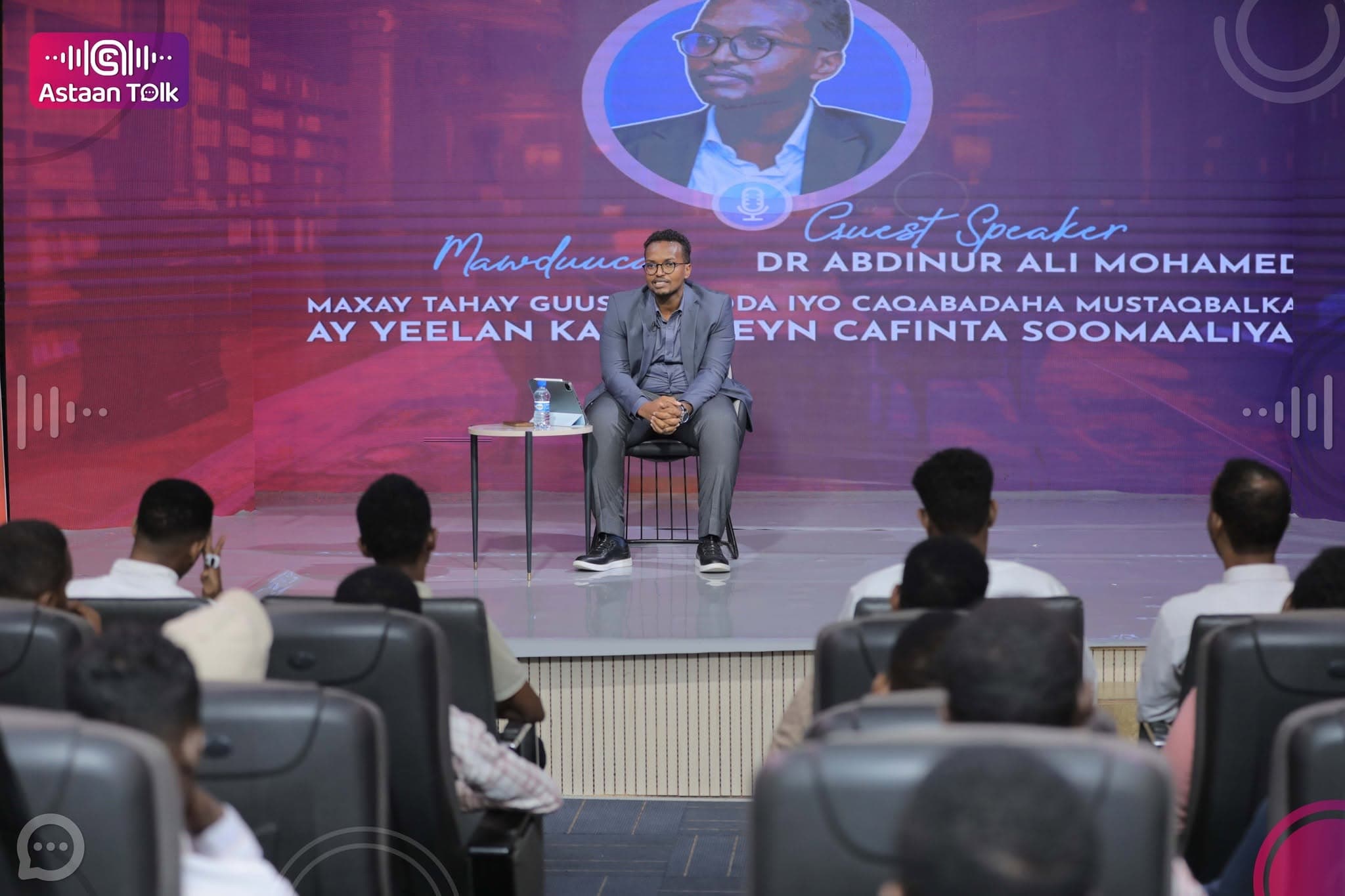 Guest speaker Dr. Abdinur Ali Mohamed seated before the audience, his name and Somali session title displayed on the purple LED screen behind him