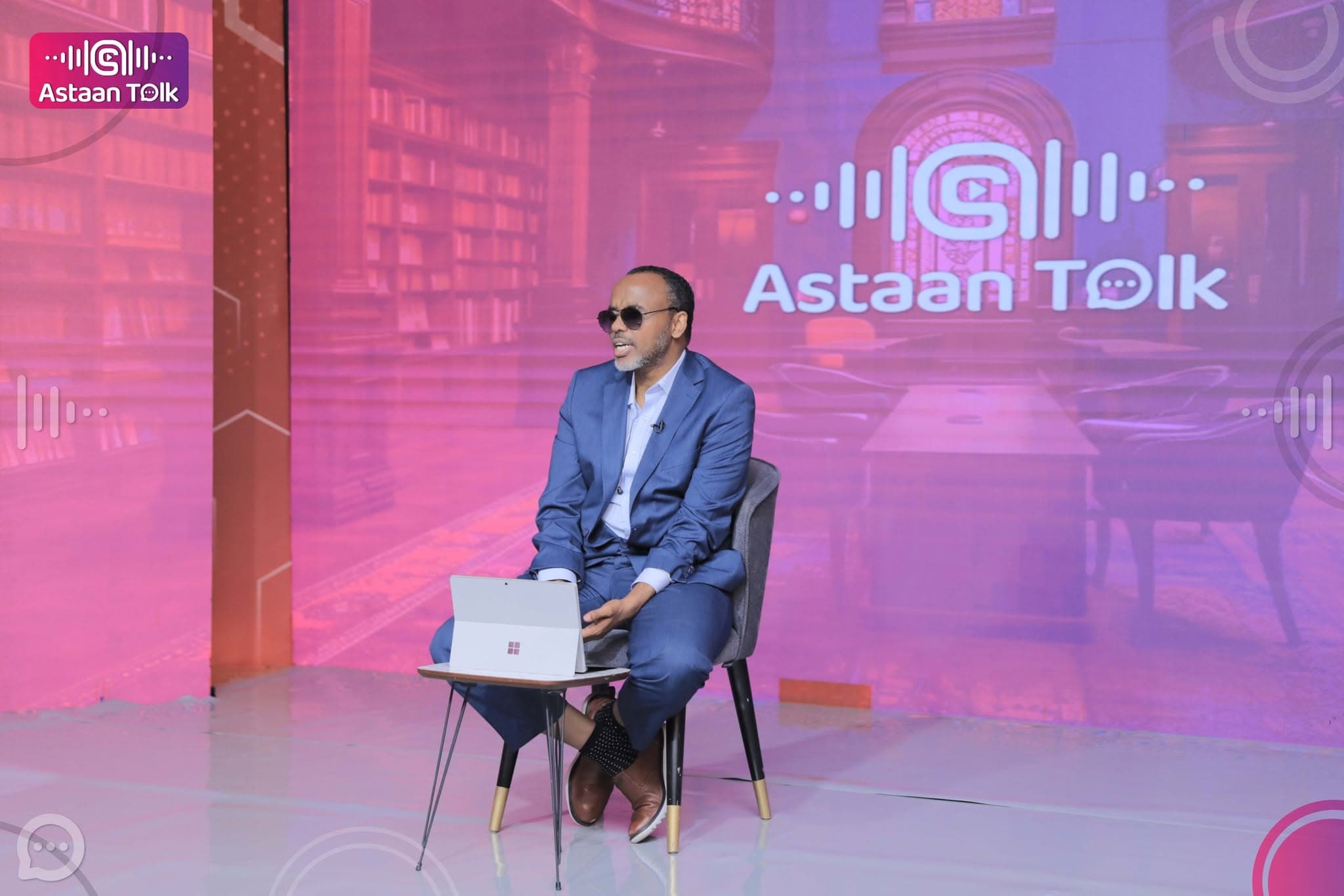 Host in a blue suit seated with a laptop on the Astaan Talk set, in front of a magenta virtual-library LED wall with the waveform wordmark