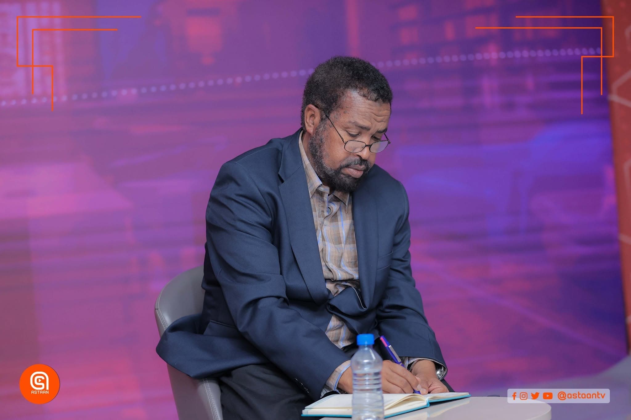 Panelist in glasses seated on stage under a purple and red wash, writing notes in a notebook