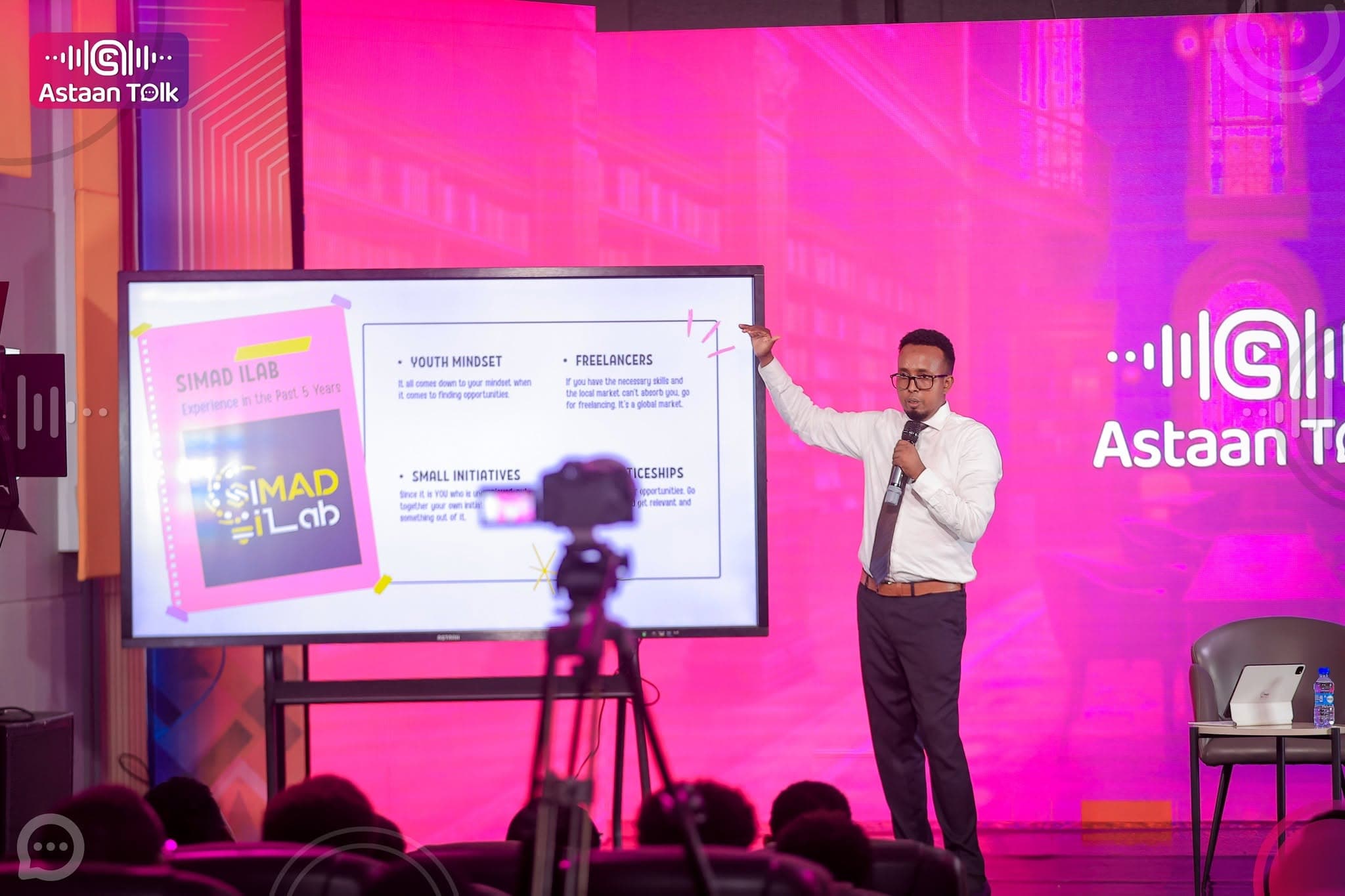 Presenter in a white shirt pointing at slides on a large screen, with the Astaan Talk waveform logo glowing on the pink stage wall beside him