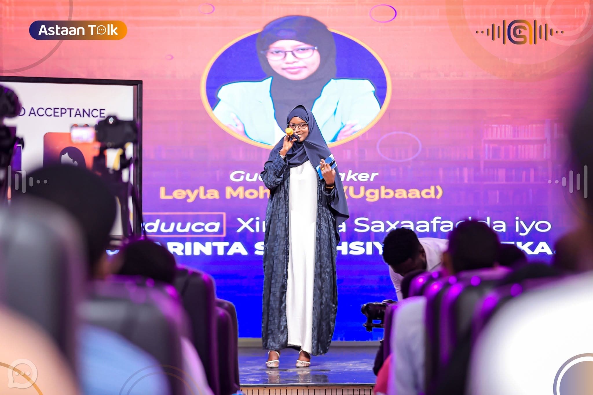 Guest speaker Leyla Ugbaad in a navy headscarf speaking into a microphone on stage before a pink LED screen announcing her session