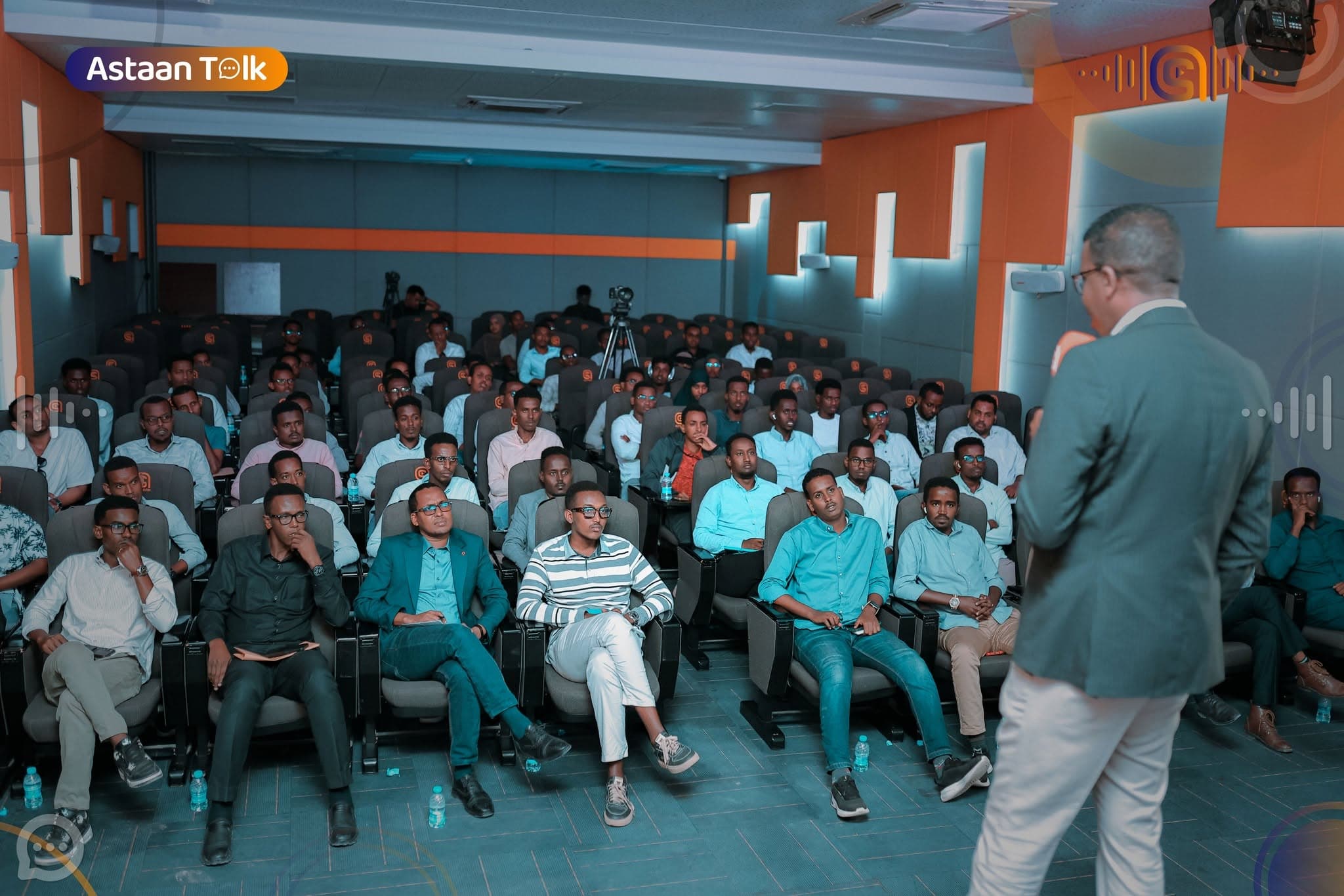 Packed auditorium of young audience members in teal-lit seats with orange wall accents, listening to an Astaan Talk speaker