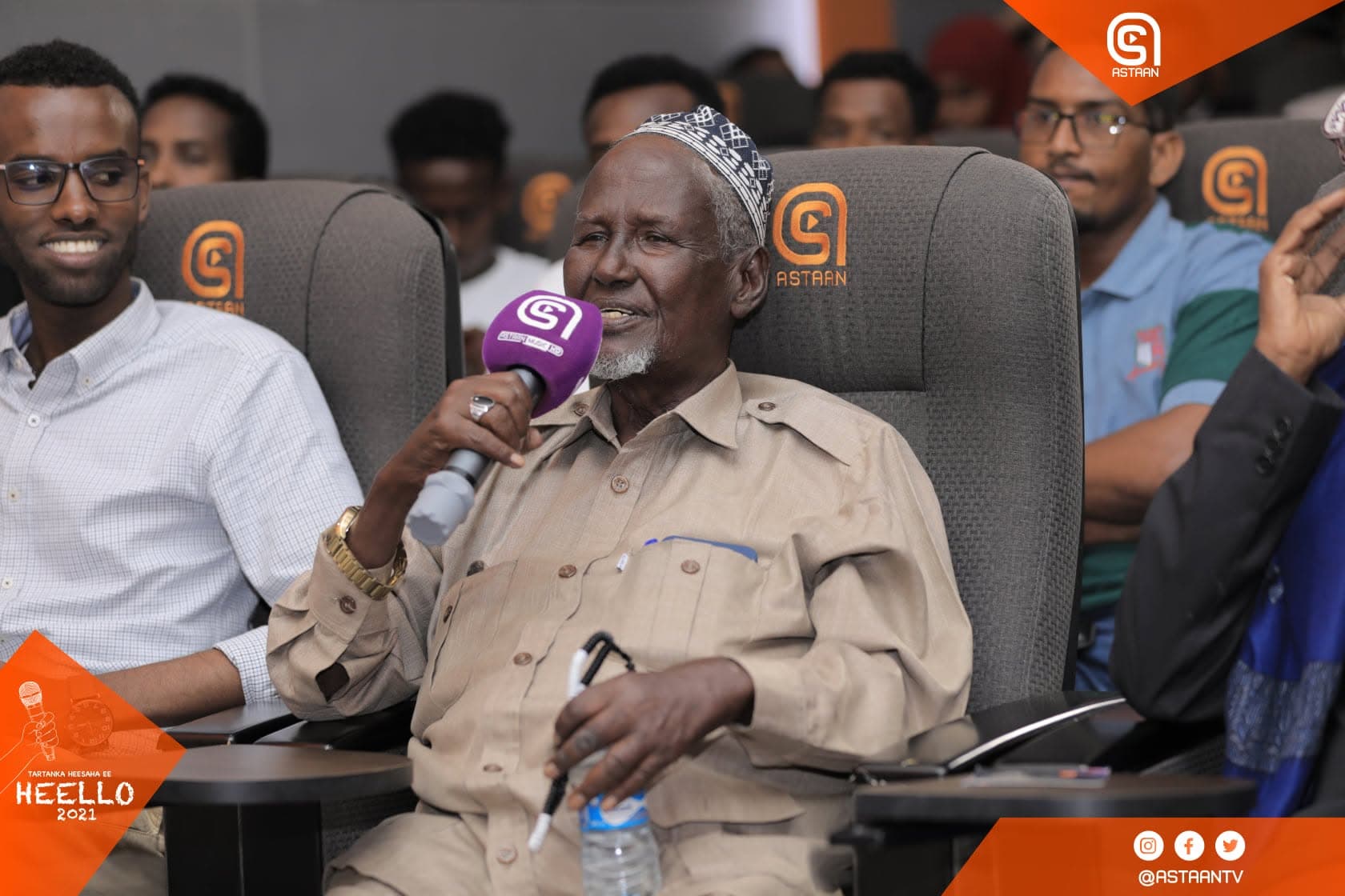Elderly audience member in an embroidered cap speaking into a purple Astaan microphone from an orange-branded studio seat among the crowd