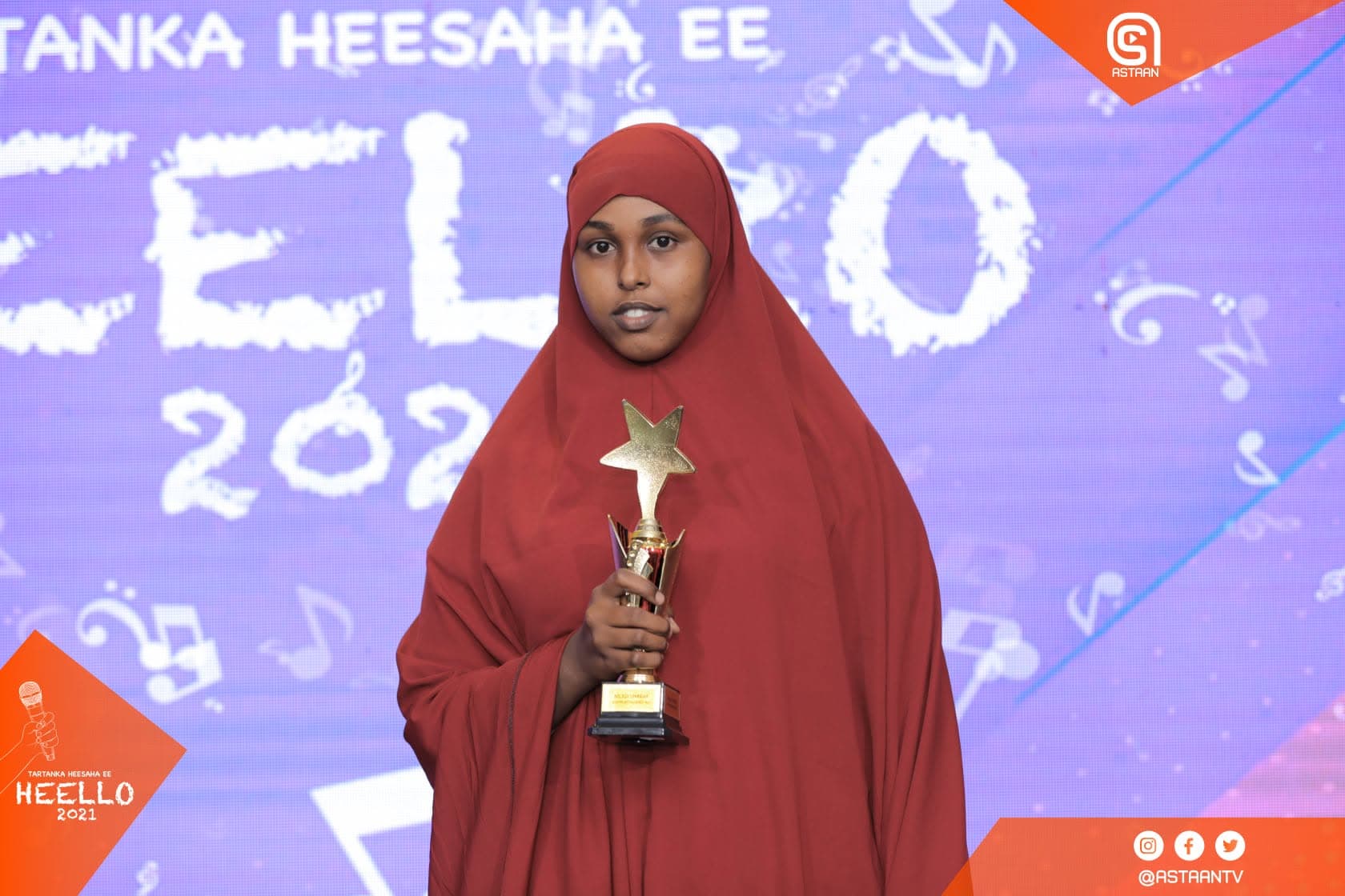 A woman in a red hijab holding a gold star trophy in front of a hand-painted Heello 2021 mural backdrop