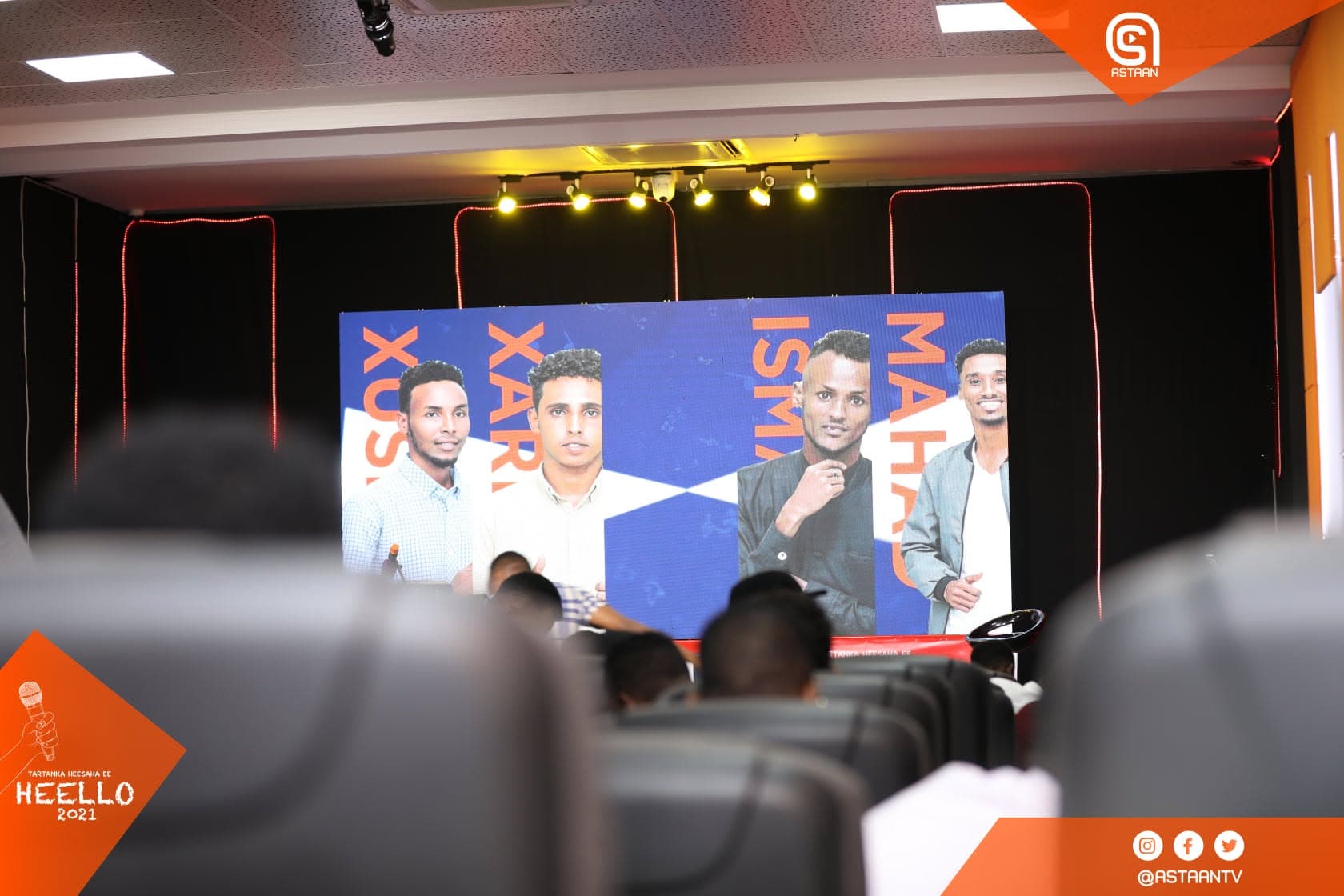 Wide studio view over audience silhouettes toward the stage screen showing contestant portrait cards on a blue graphic