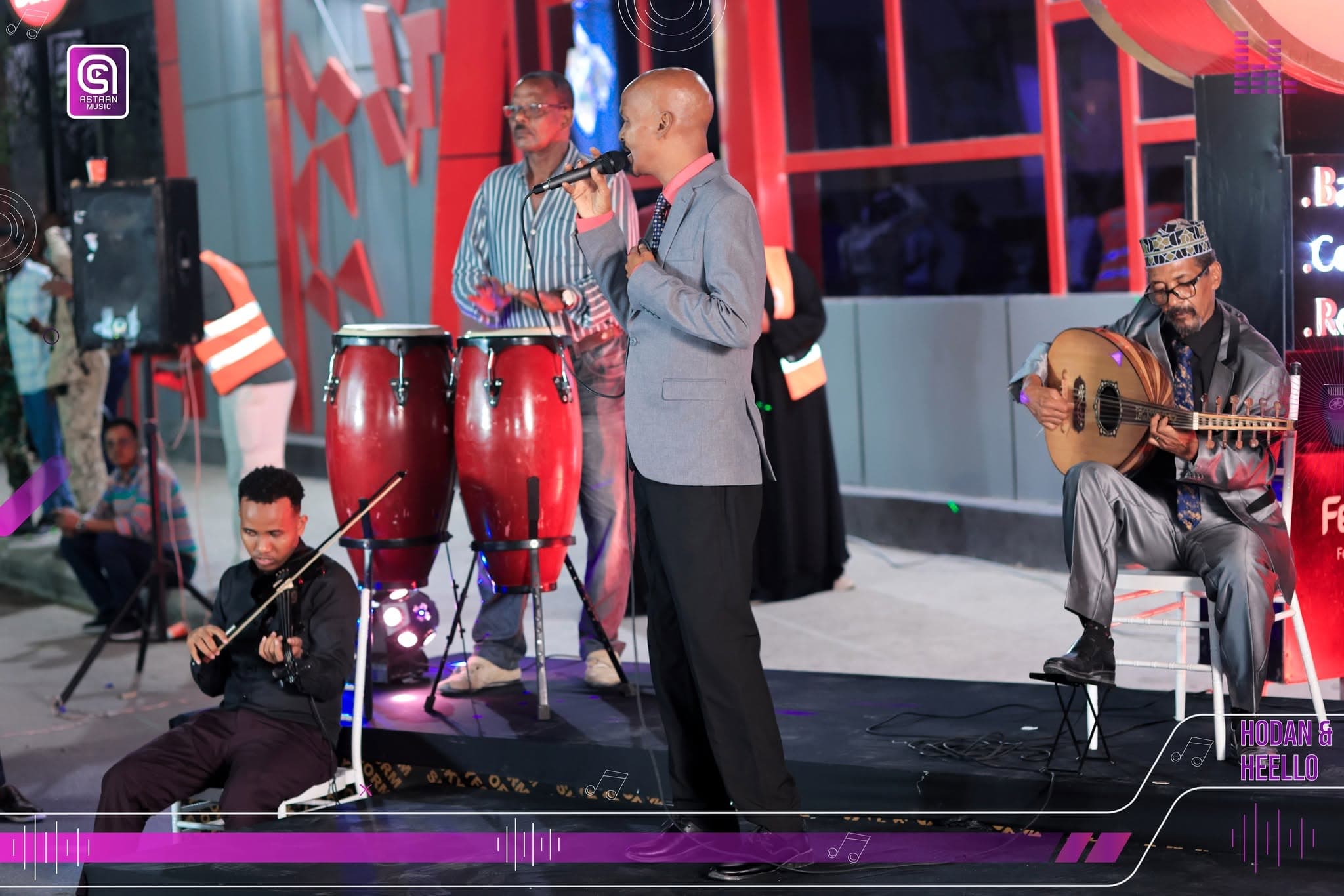 Musica Band on the street stage at night: a seated violinist, red conga drums, the suited lead singer at the microphone and a seated oud player against a red-lit wall