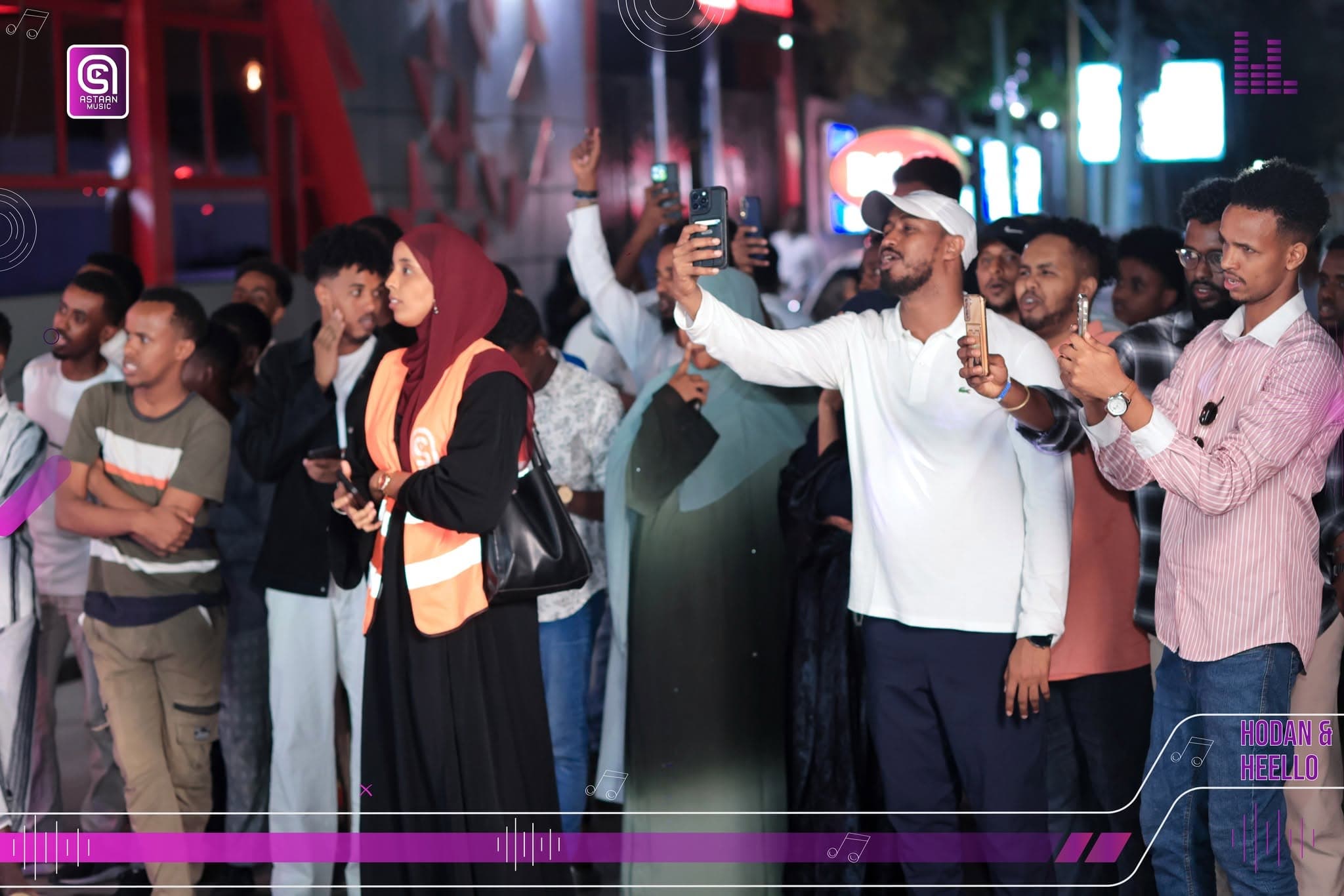 Night-time audience filming the performance with raised phones; a woman in a maroon hijab and orange safety vest stands in the foreground while a man in a white cap takes a selfie
