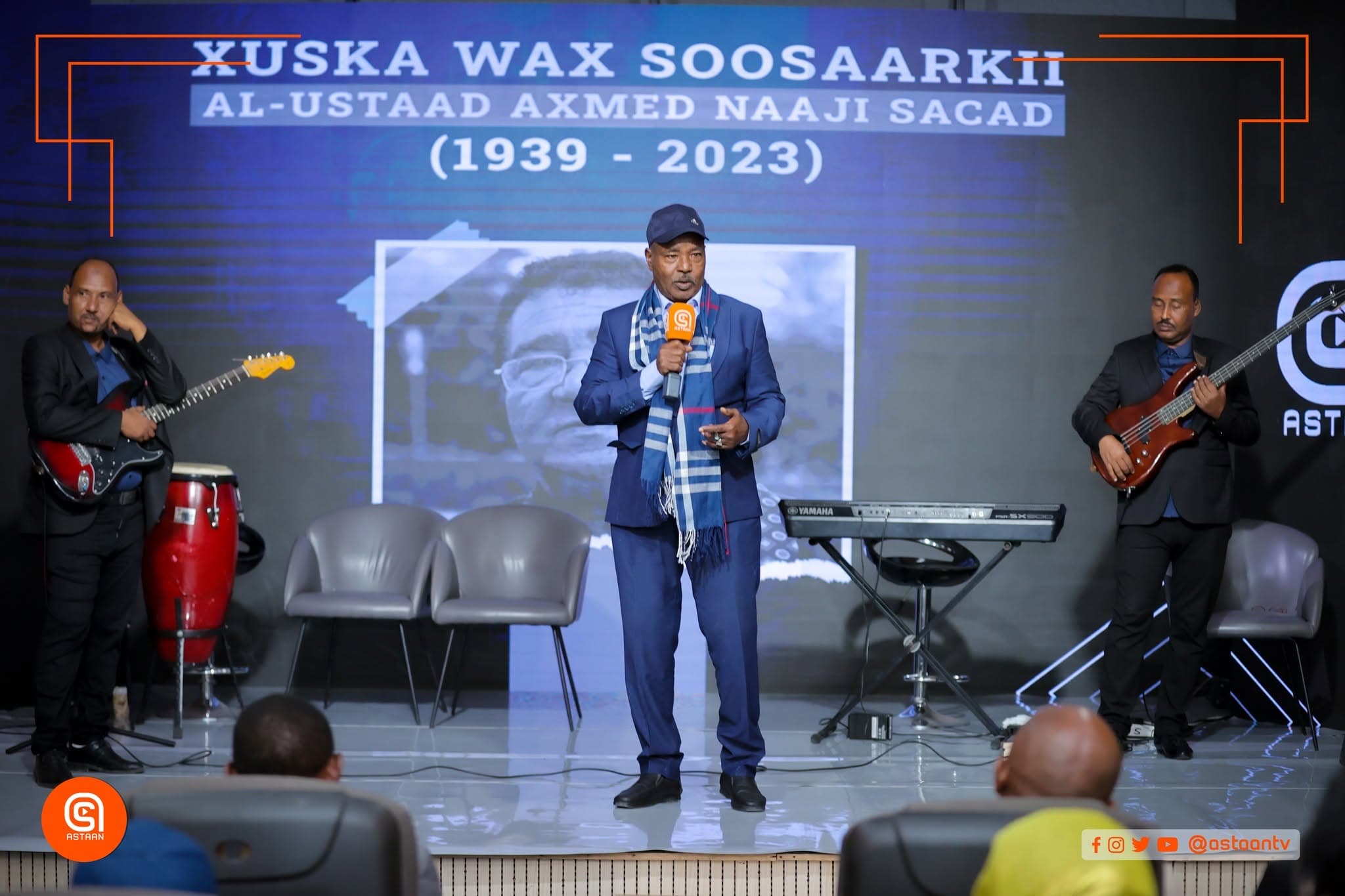 A singer in a blue suit and checked scarf performs at center stage with the band at the Axmed Naaji Sacad commemoration, Astaan headquarters.