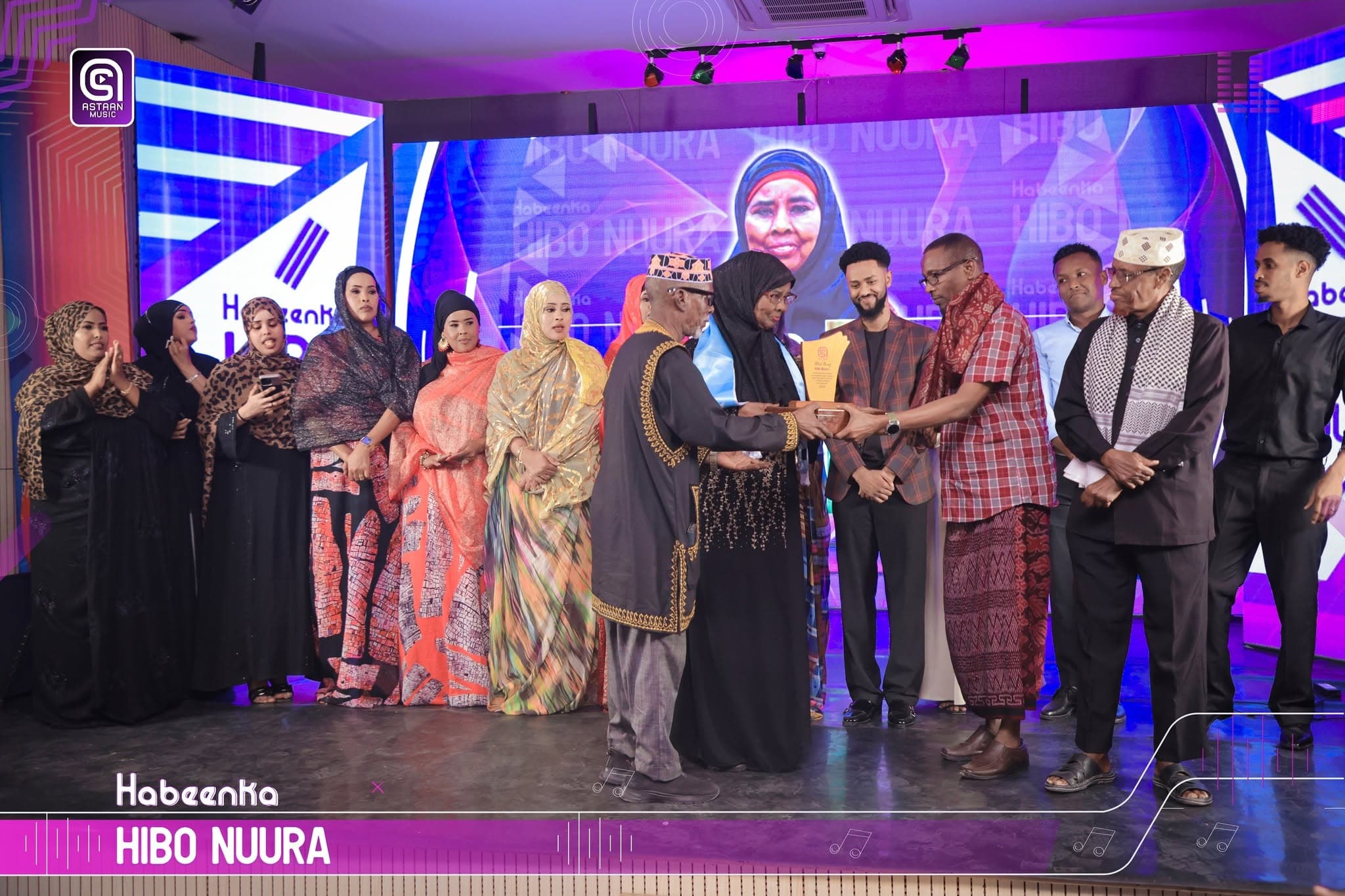 Award handover at the Habeenka Hibo Nuura tribute: a line of guests and performers in traditional Somali dress on stage, with Hibo Nuura's face on the purple LED wall behind them.