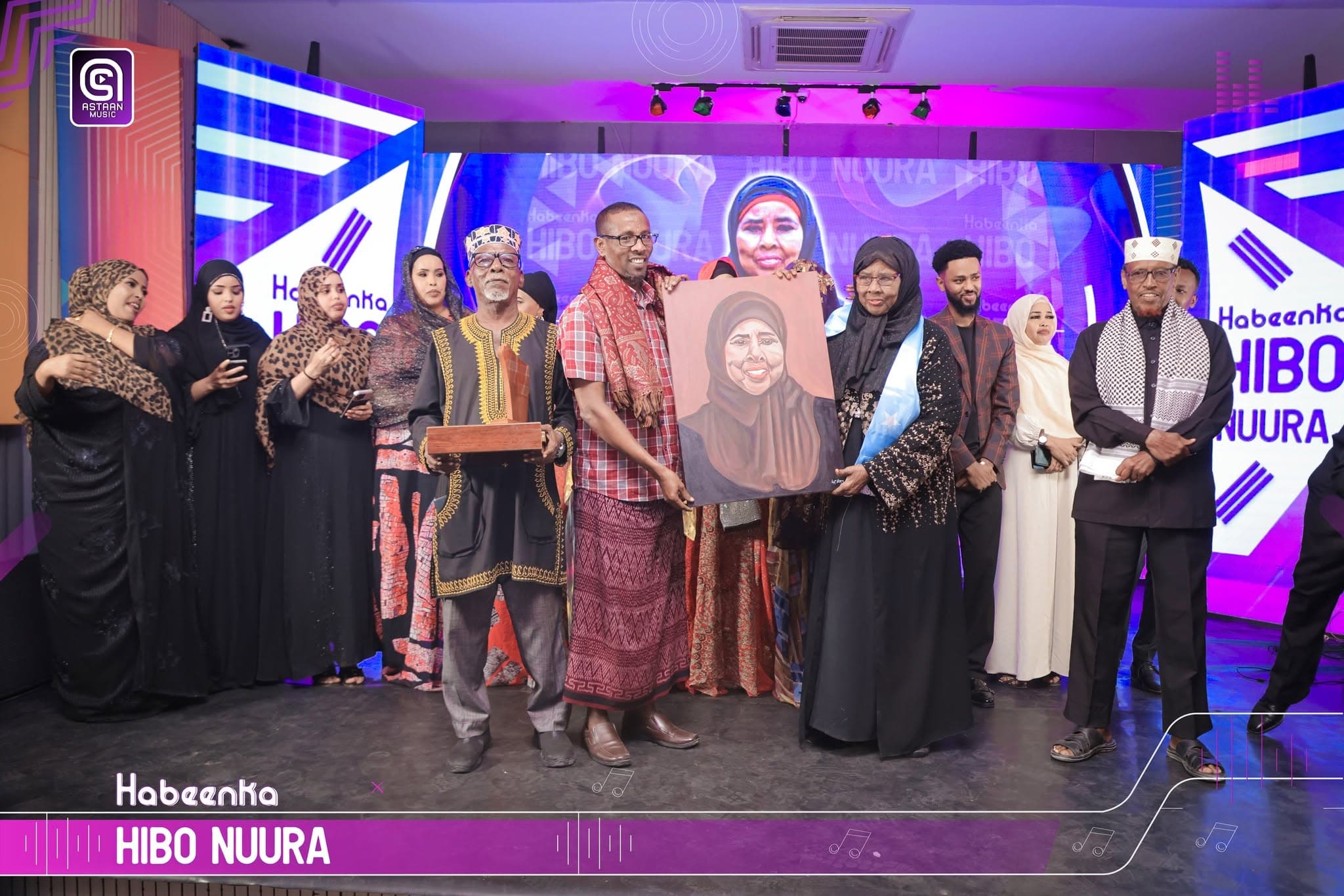 The ensemble of the Habeenka Hibo Nuura tribute on stage, raising framed portrait paintings of the singer before a purple LED backdrop.