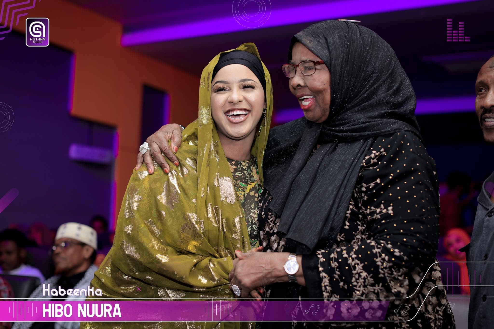 A young artist in a gold-embroidered shawl embraces Hibo Nuura, both laughing under magenta and purple stage light.