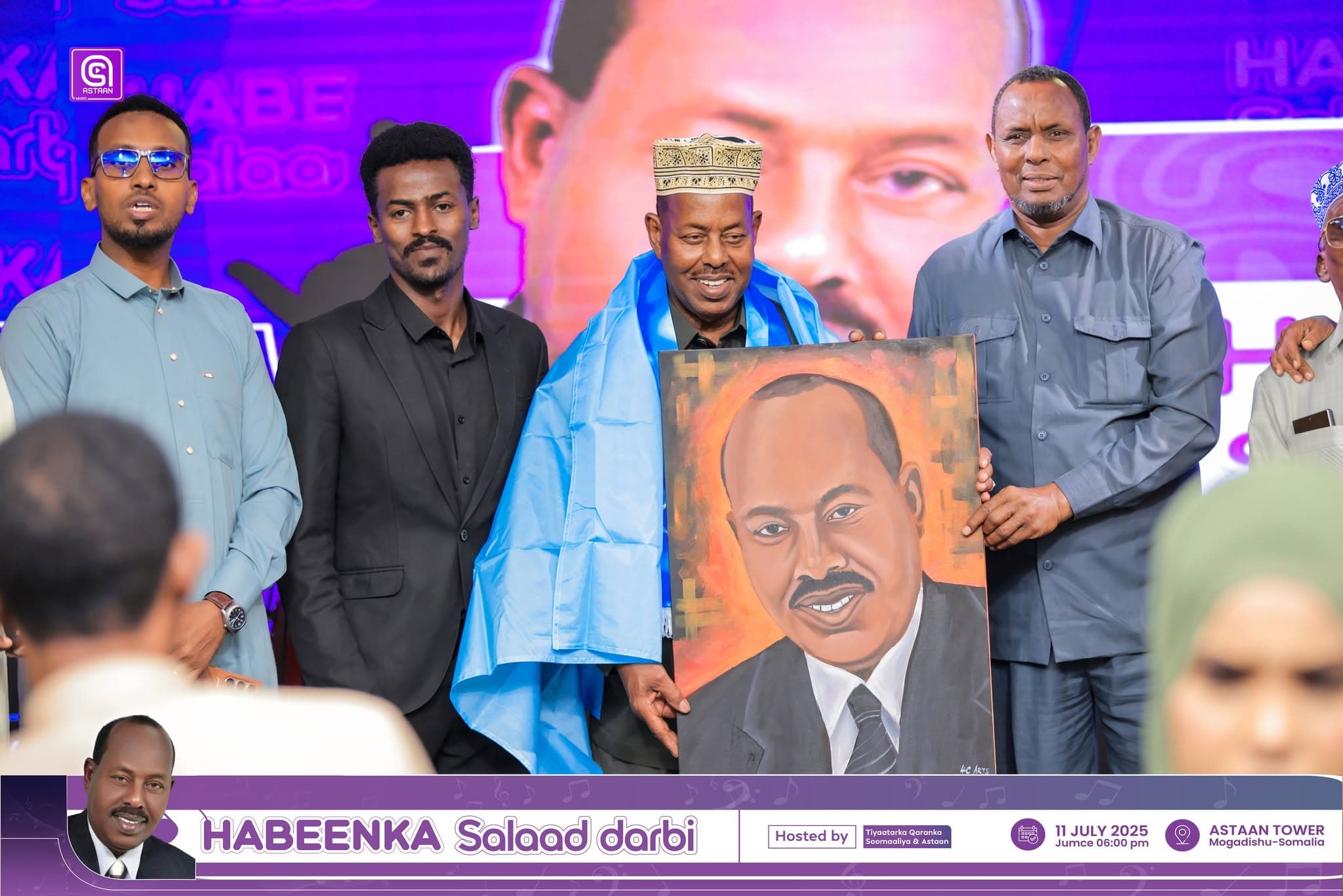 Five men across the Habeenka Salaad Darbi stage; at center, the honoree in blue ceremonial drape and crown-style koofiyad holds the framed portrait painting of Salaad Darbi.