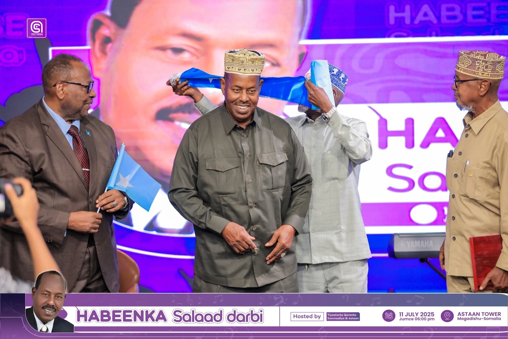 A blue ceremonial drape is placed over the shoulders of an honoree wearing a crown-style koofiyad on the Habeenka Salaad Darbi stage, flanked by elders holding Somali flags.