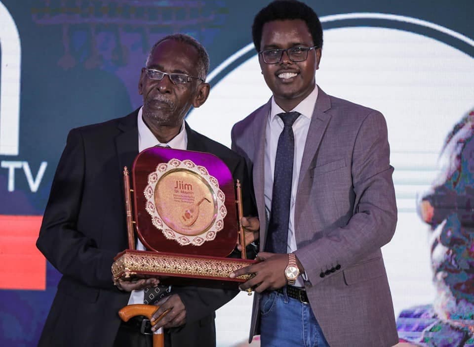 Elderly Muxiyadiin Sheekh Muumin (Jiim), in glasses and a dark suit, receives a circular gold award plaque engraved with his name from a smiling presenter in a grey suit.