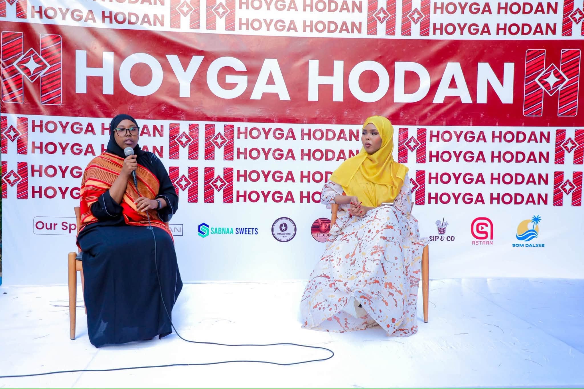 Two women seated in conversation on stage in front of a red-and-white Hoyga Hodan sponsor wall