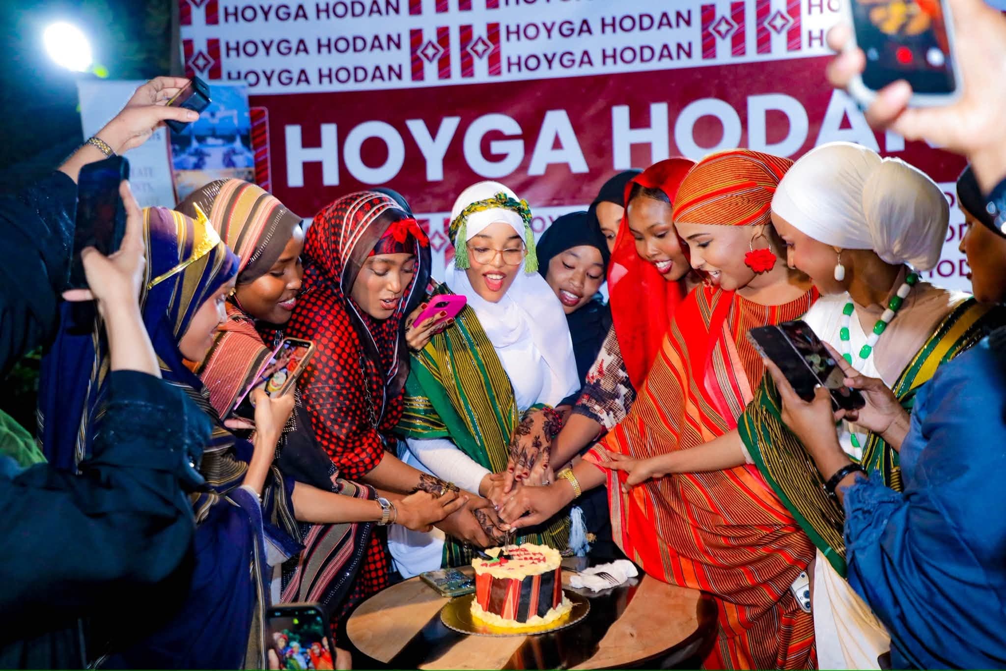 Women in colourful guntiino gather around a cake, phones raised, at a Hoyga Hodan celebration