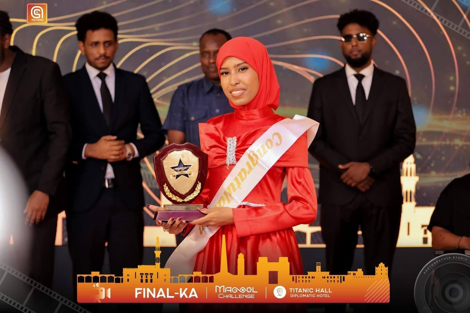 The Magool Challenge winner in a bright red satin dress and red hijab, wearing a gold Congratulations sash and holding a shield-shaped trophy, flanked by men in dark suits on a stage lit with golden light-trails