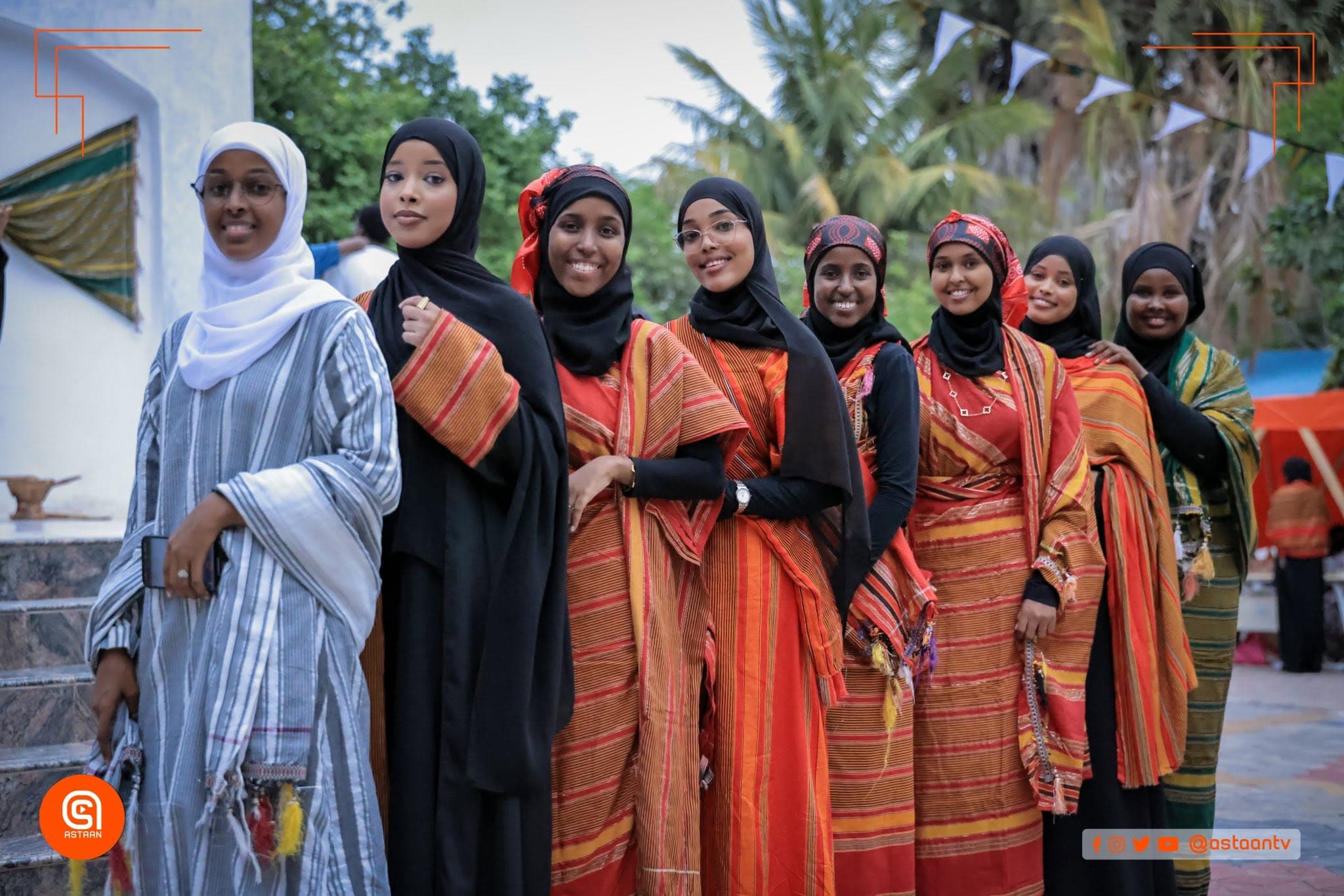 Smiling Magool Challenge contestants standing together outdoors in bright orange, green, and patterned traditional Somali dress with black, white, and patterned hijabs, trees behind them