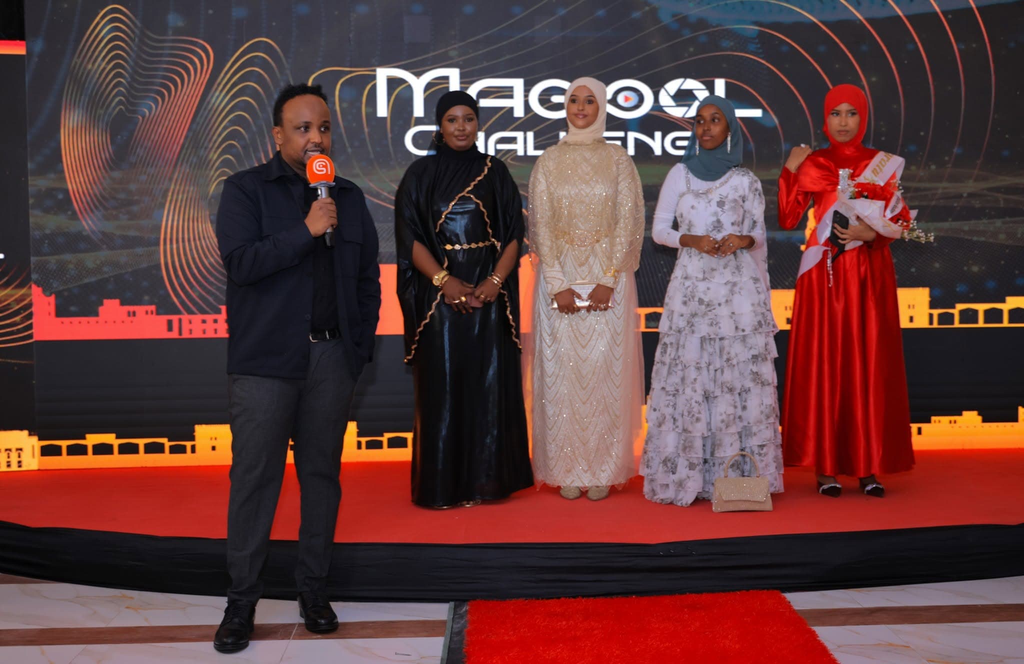 The Magool Challenge grand finale: a host in a dark suit speaks into a microphone beside four finalists in elegant gowns, in front of the glowing Magool Challenge logotype on an LED stage wall