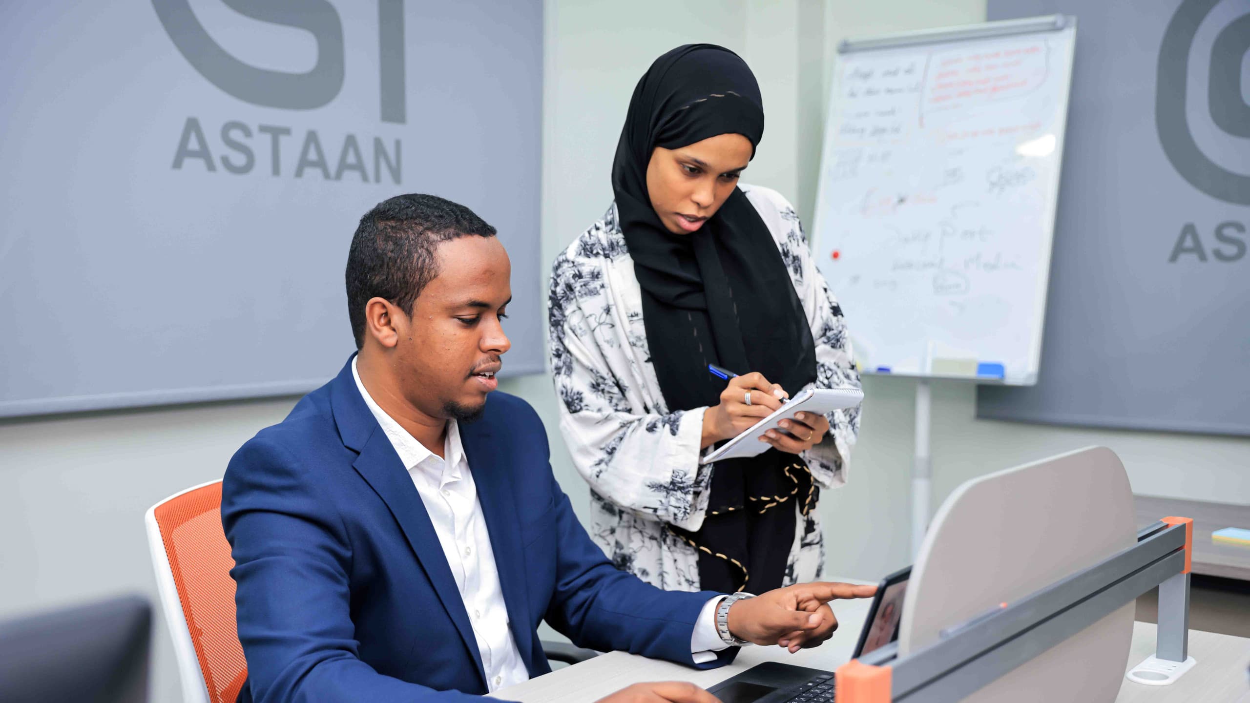 Two Astaan strategists planning a campaign at a laptop, one taking notes beside a whiteboard